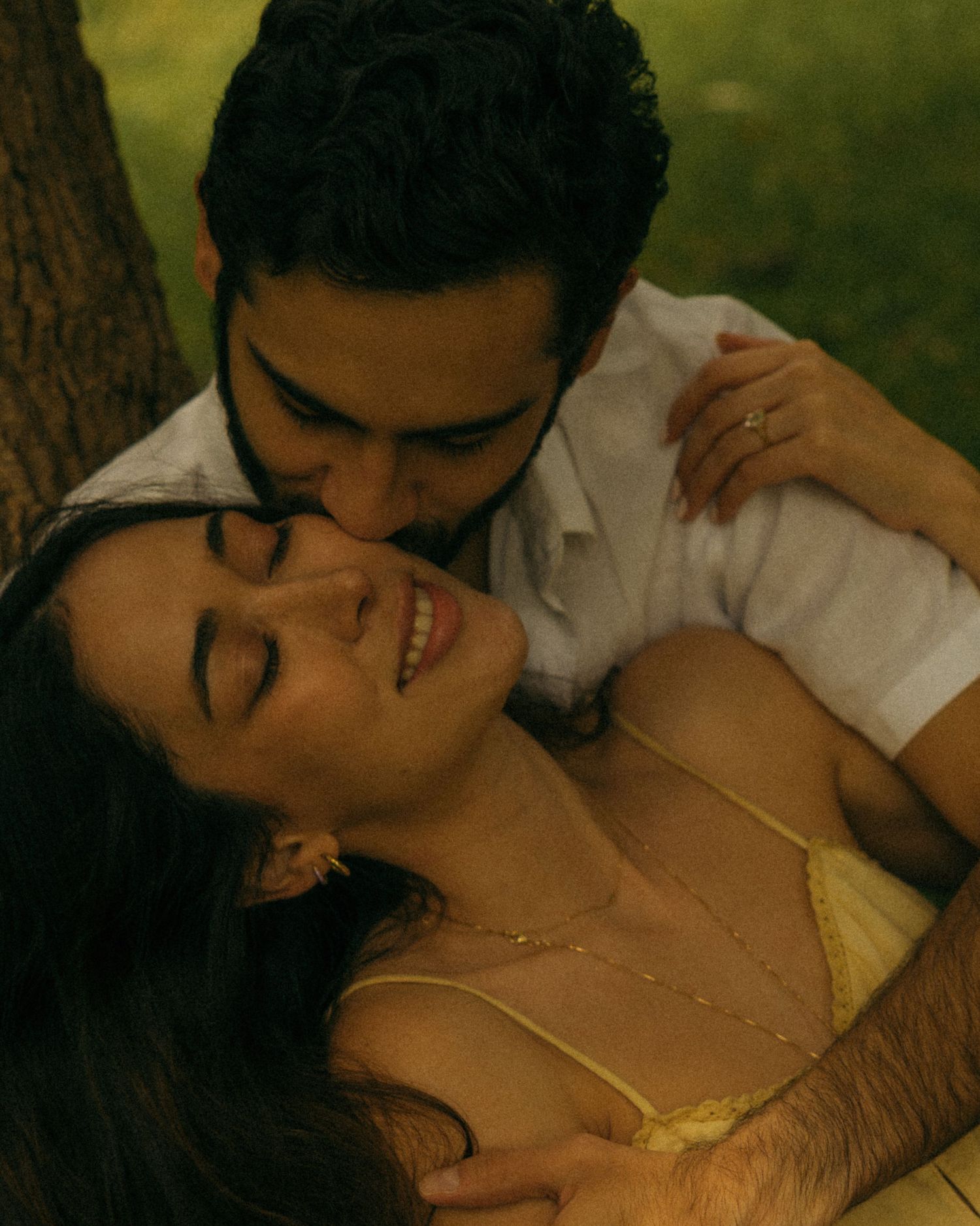 A couple shares a romantic moment outdoors, with a man kissing a smiling woman near a tree.
