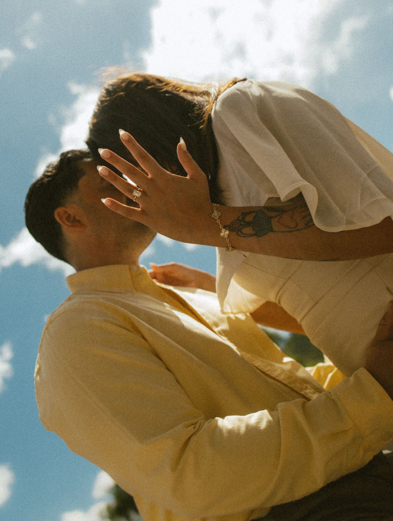 Couple in an embrace and kiss under a bright blue sky, her hand with a ring on his face, wearing a yellow shirt and white top.