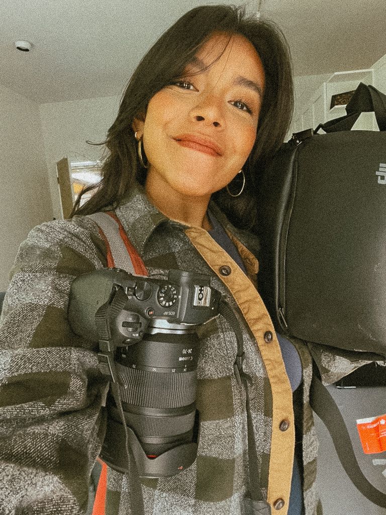 Woman taking a mirror selfie wearing a plaid shirt with a Canon DSLR camera around her neck.