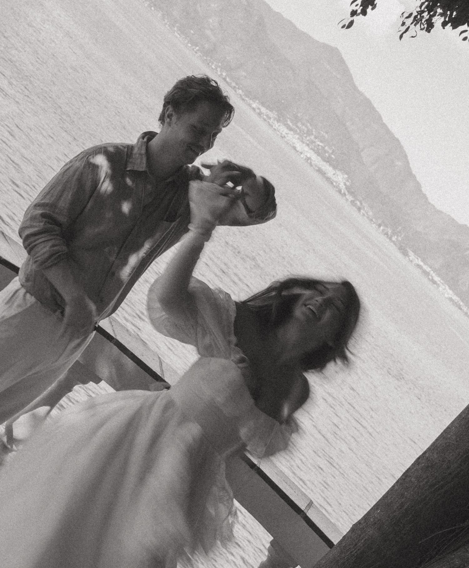 Black and white photo of couple dancing on dock with mountain lake backdrop.