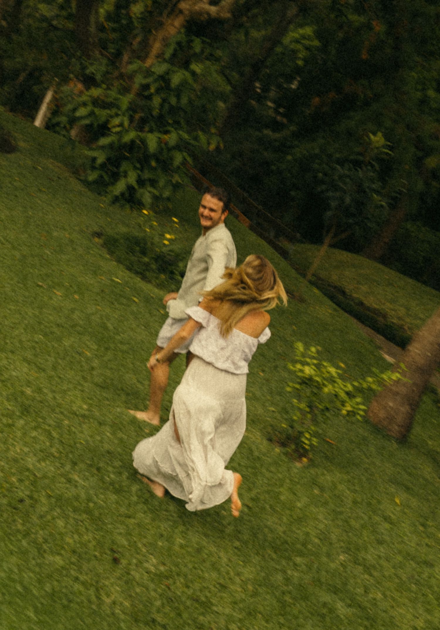 Couple dancing and spinning joyfully on a lush green hillside surrounded by tropical trees.