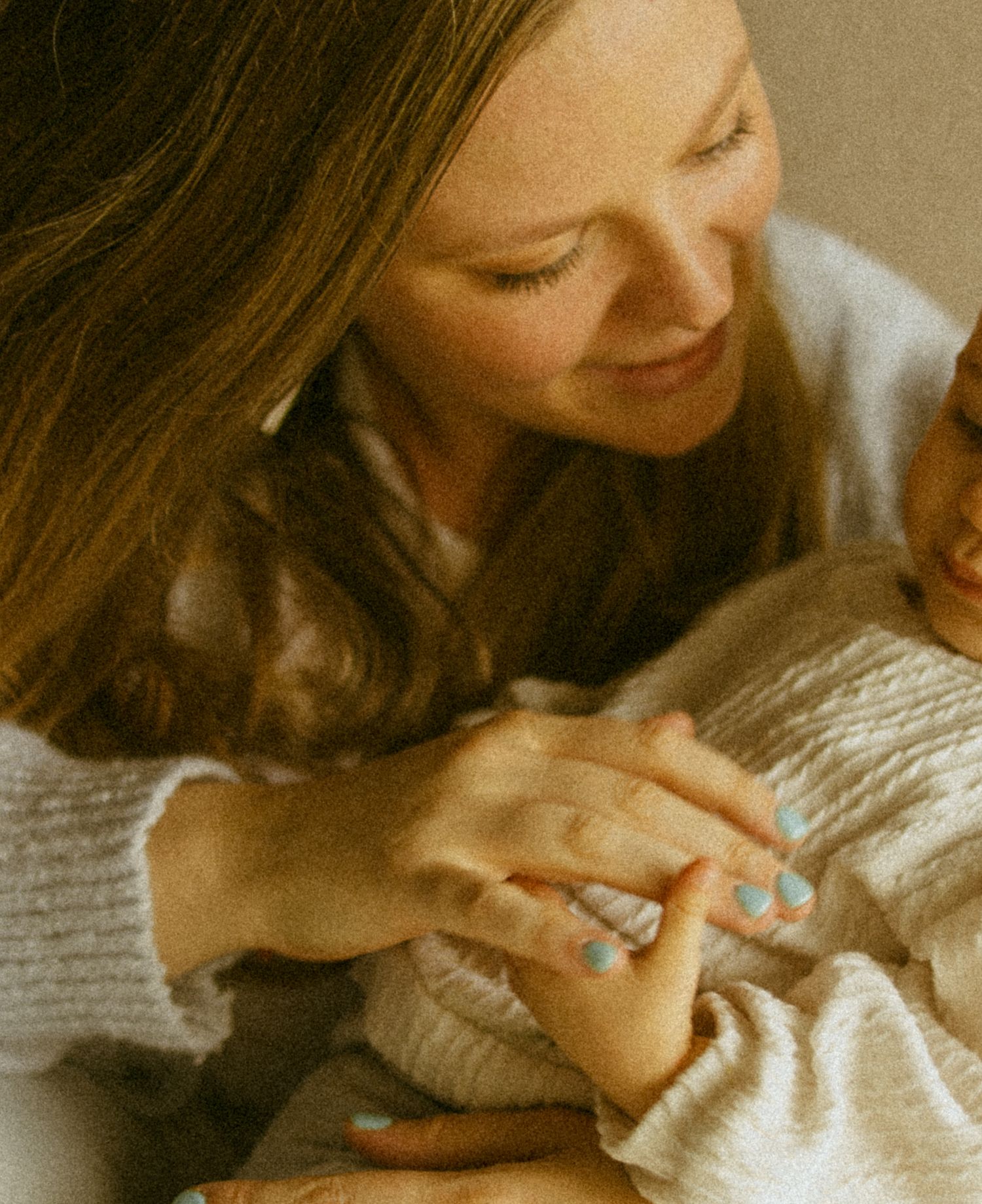 A smiling mother with long hair tenderly holds her newborn baby, wearing a cozy white sweater and blue nail polish.