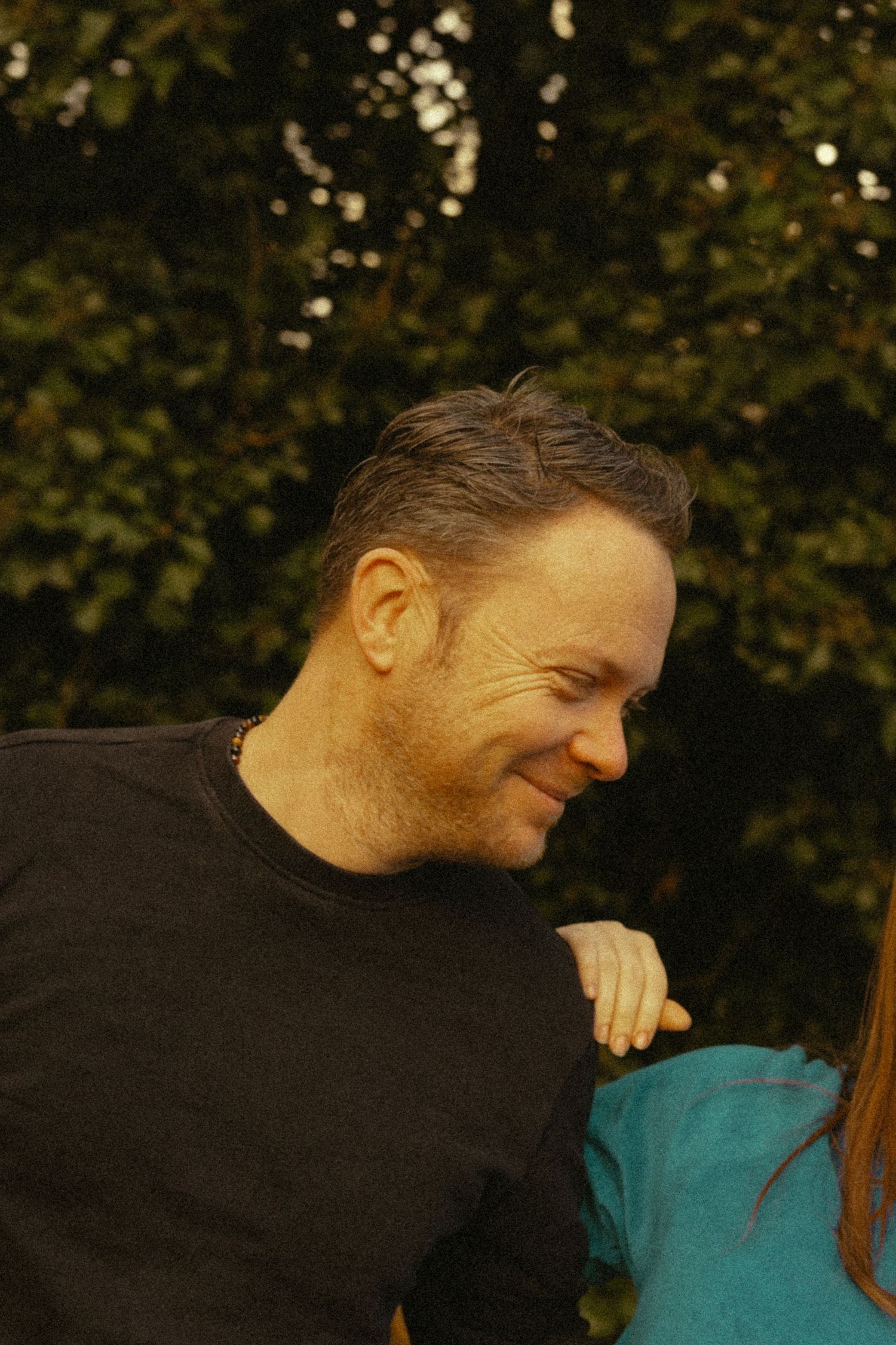 A smiling middle-aged man in a black t-shirt with face paint, standing outdoors near green trees.