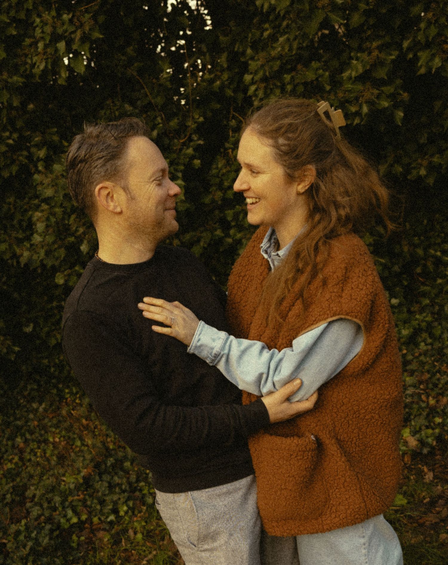A smiling couple embraces outdoors in front of lush green foliage, woman in rust knit vest and man in black sweater.