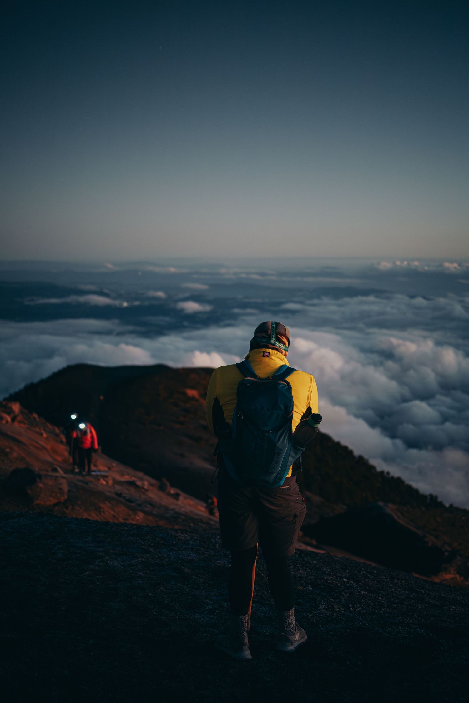 Hiker in yellow jacket stands above clouds on volcanic mountain ridge at dusk with another climber visible below.