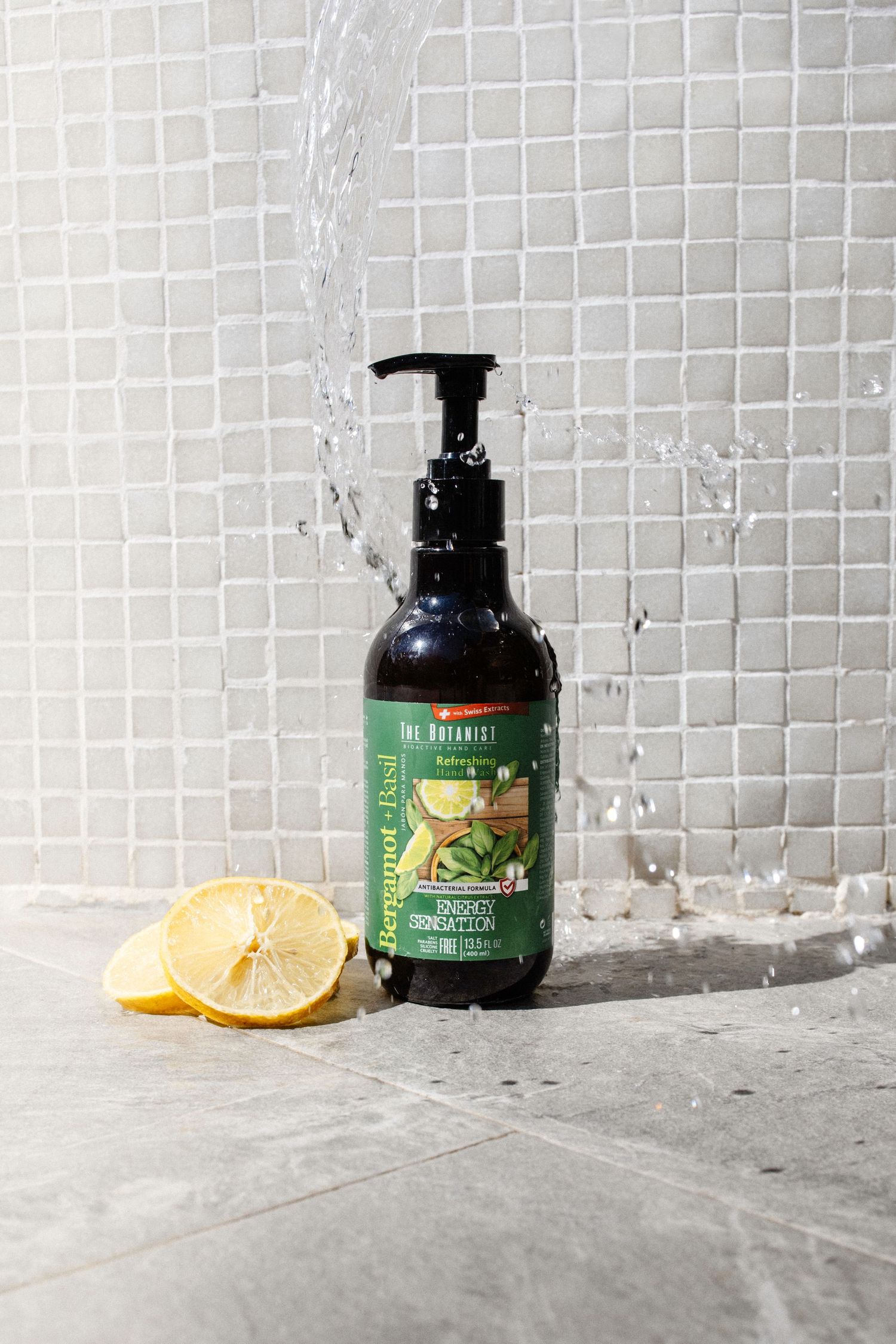 The Botanist Bergamot & Basil body lotion pump bottle beside a lemon half, with water splashing on white tile.
