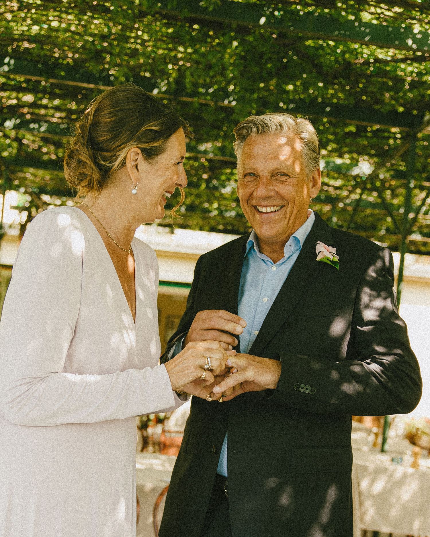 A bride in white dress and groom in dark suit laugh joyfully while exchanging rings outdoors under green pergola.