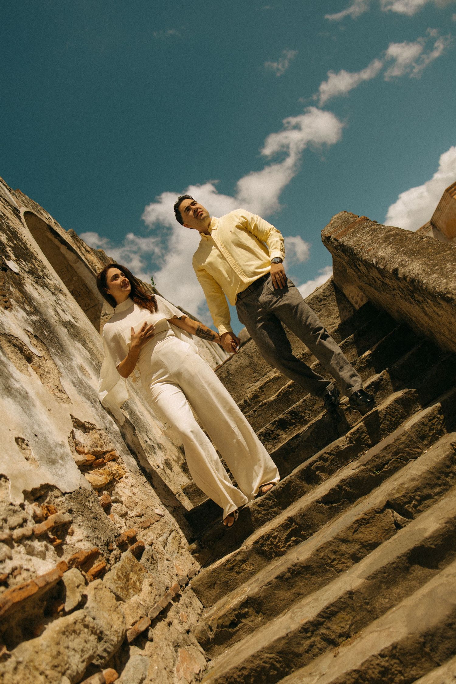 Couple descending ancient stone steps under blue sky, woman in white outfit and man in yellow shirt at historic ruins.
