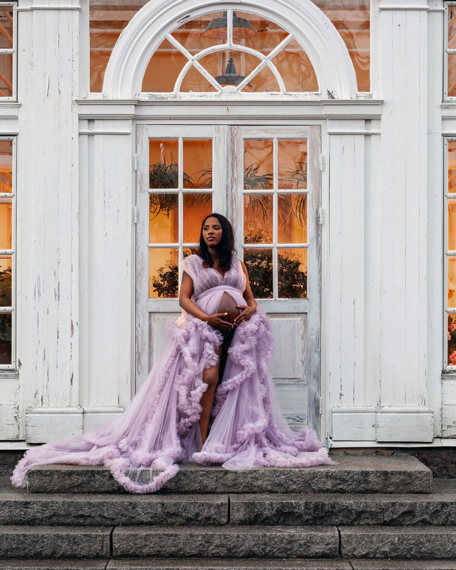 A pregnant woman in a lavender ruffled tulle gown poses on steps before a white vintage building door.