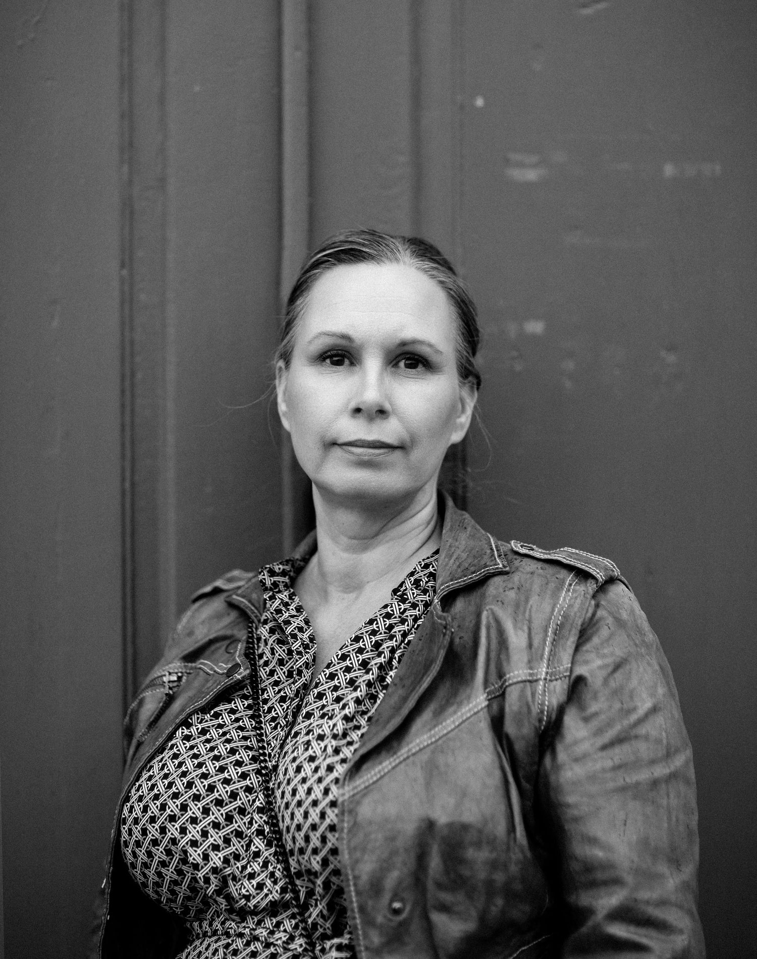 Black and white portrait of woman in leather jacket leaning against dark door outdoors.