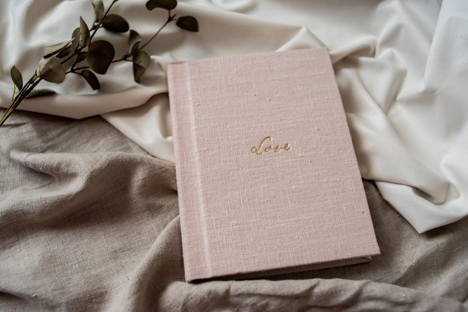 Elegant blush pink linen hardcover journal with gold script lettering resting on soft white fabric.