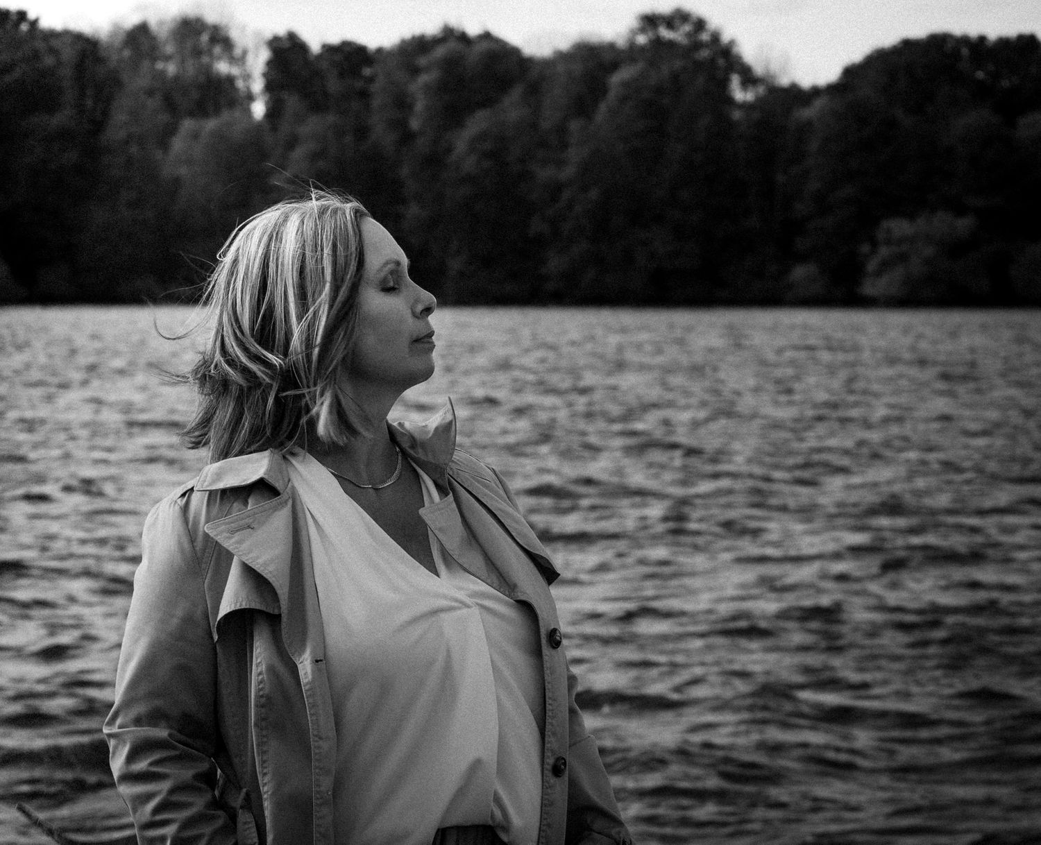 Black and white portrait of blonde pregnant woman in trench coat standing by a lake.