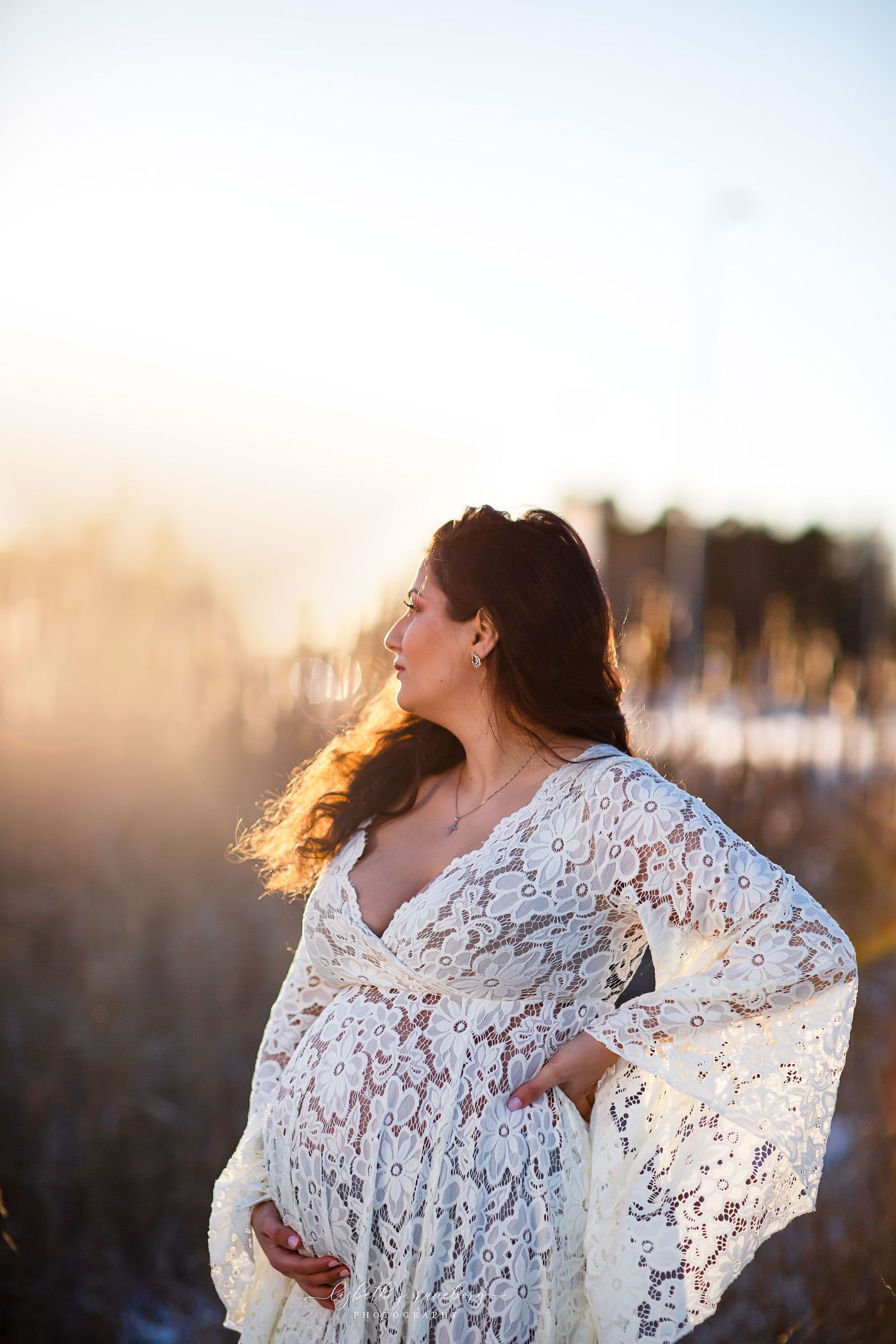 Pregnant woman in white lace dress standing in golden sunset light in a winter field.