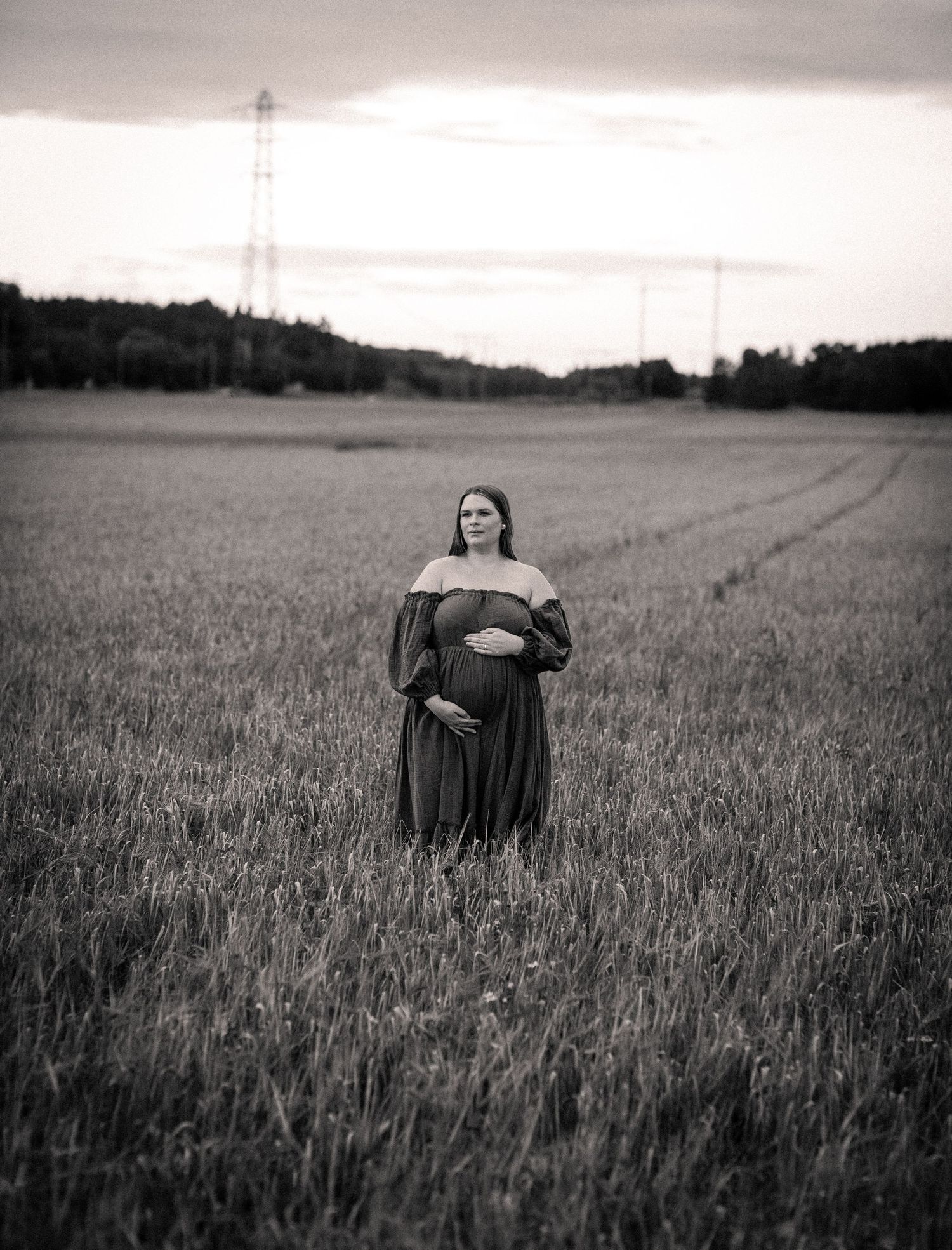 Black and white maternity portrait of woman in off-shoulder dress standing in open field at sunset.