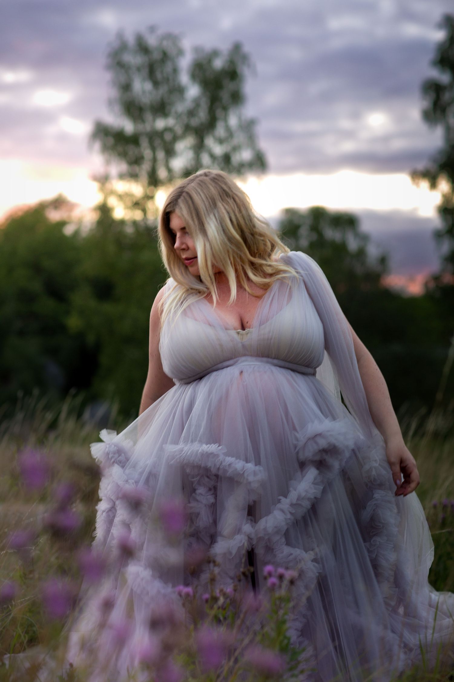 Pregnant blonde woman in flowing lavender tulle gown posing in wildflower field at dusk.