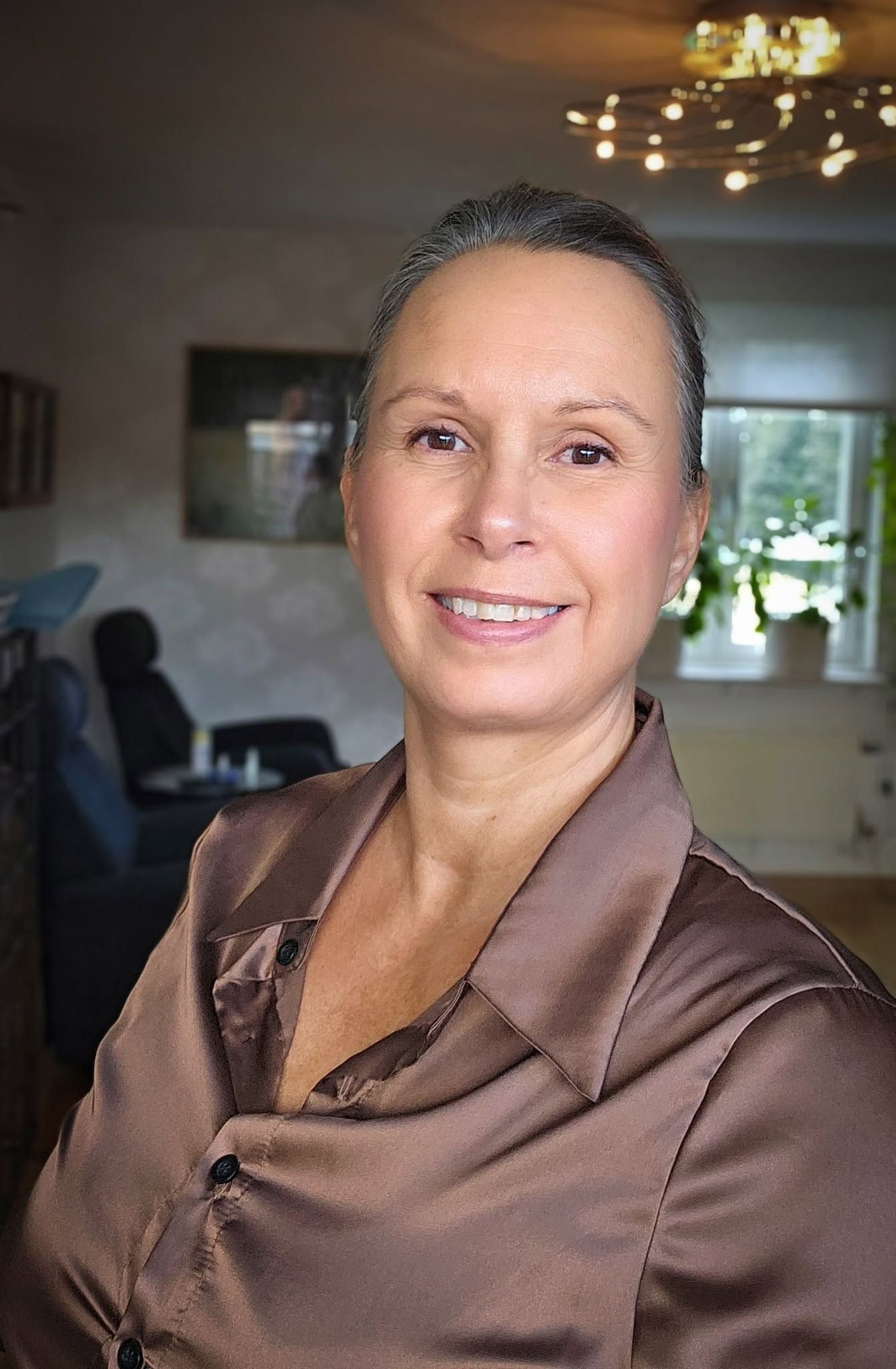 Elegant older woman with gray hair smiling in a brown satin blouse inside a warmly lit room.