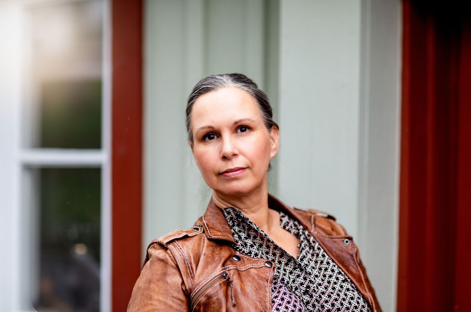Woman in brown leather jacket with patterned blouse poses outdoors near red wooden building.