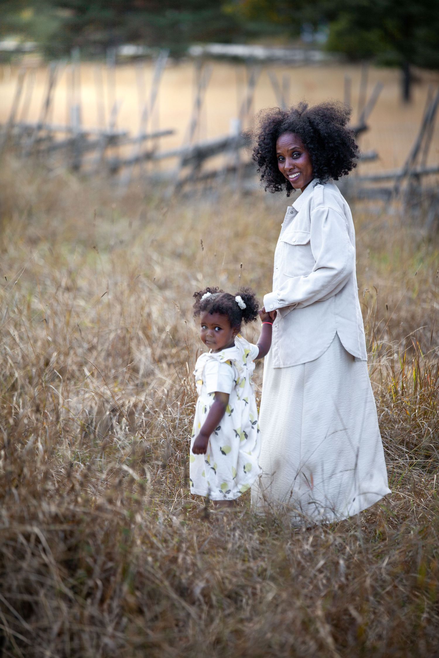 A smiling Black mother in a beige outfit holds her young daughter's hand in a golden field near a fence.