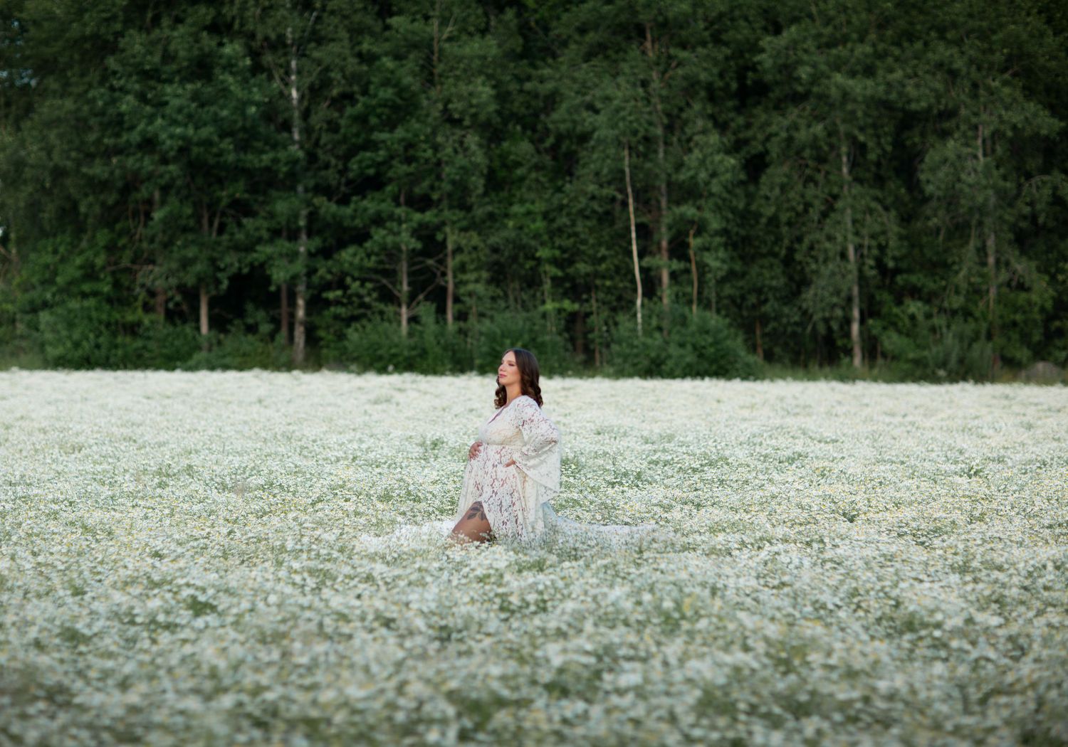 Woman in floral dress standing in vast white wildflower meadow with green forest backdrop.