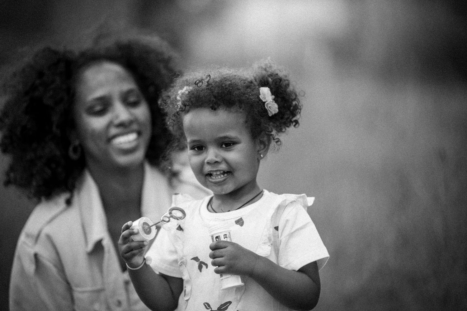 Black and white portrait of a young Black girl holding a bubble wand with her smiling mother behind her.