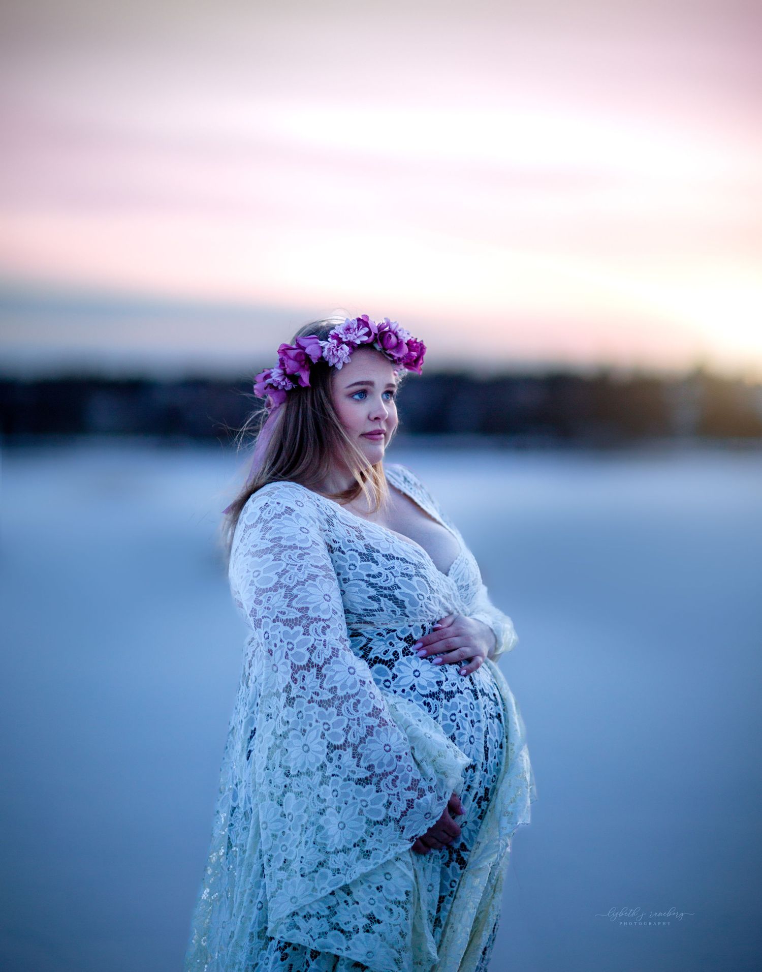 A pregnant woman wearing a floral crown and white lace shawl poses gracefully in a snowy twilight setting.