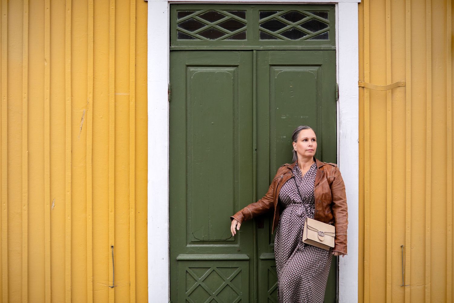 Woman in brown leather jacket and floral dress leans against green door on yellow building.