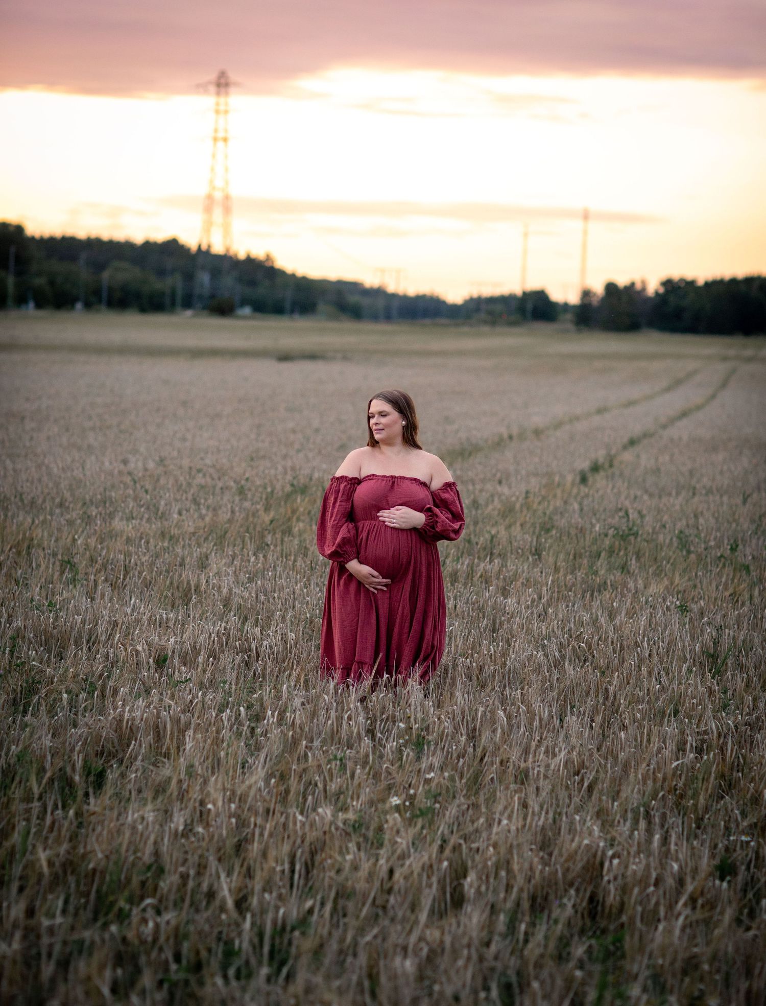 Pregnant woman in red off-shoulder gown standing in golden field at sunset with power lines in background.