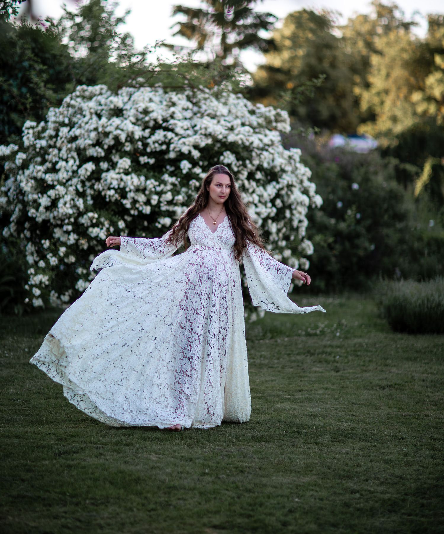 Woman in white lace boho wedding dress twirling on green lawn near white flowering shrub.