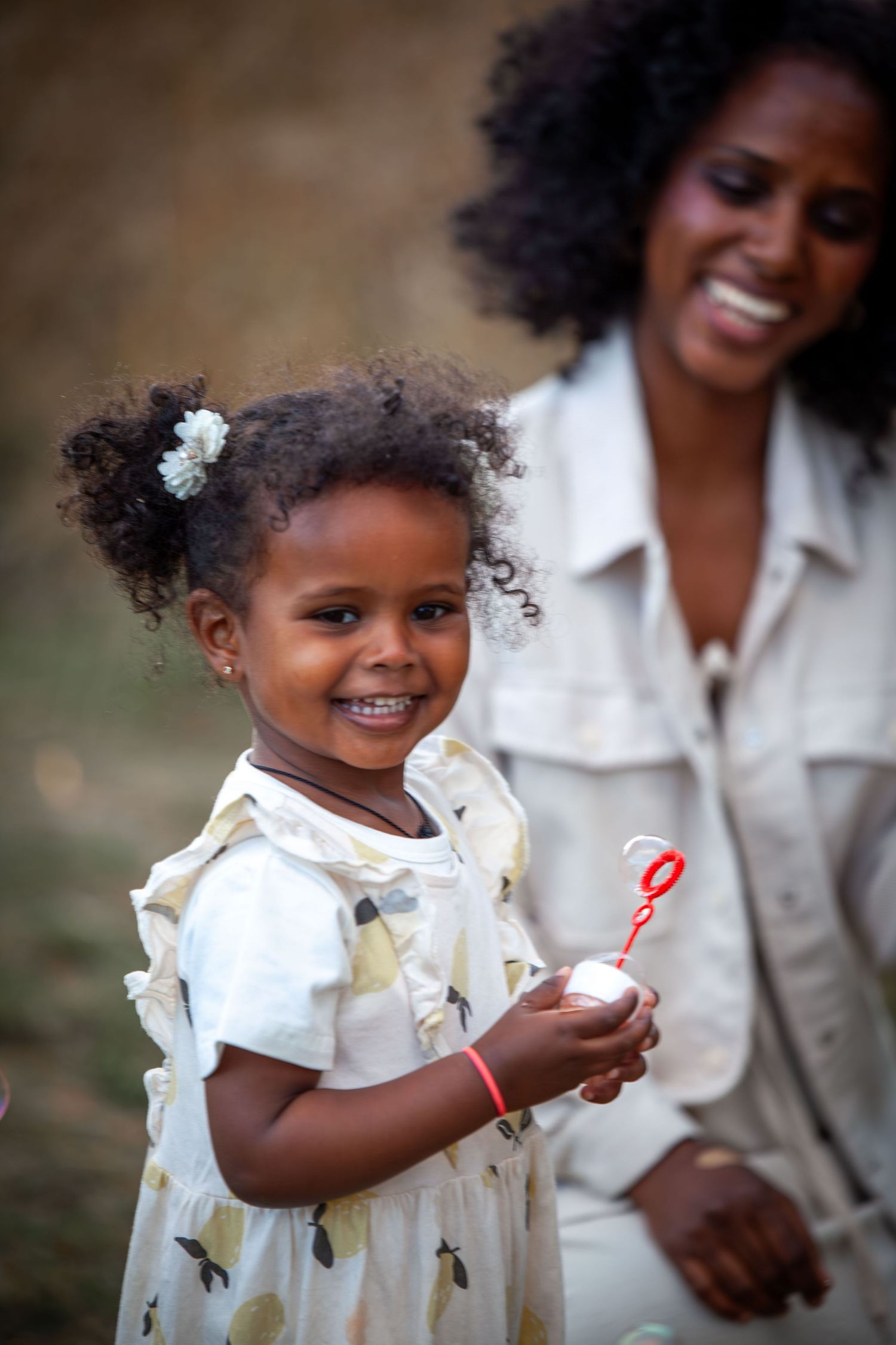 A joyful young Black girl in a floral dress blows bubbles while her smiling mother watches outdoors.
