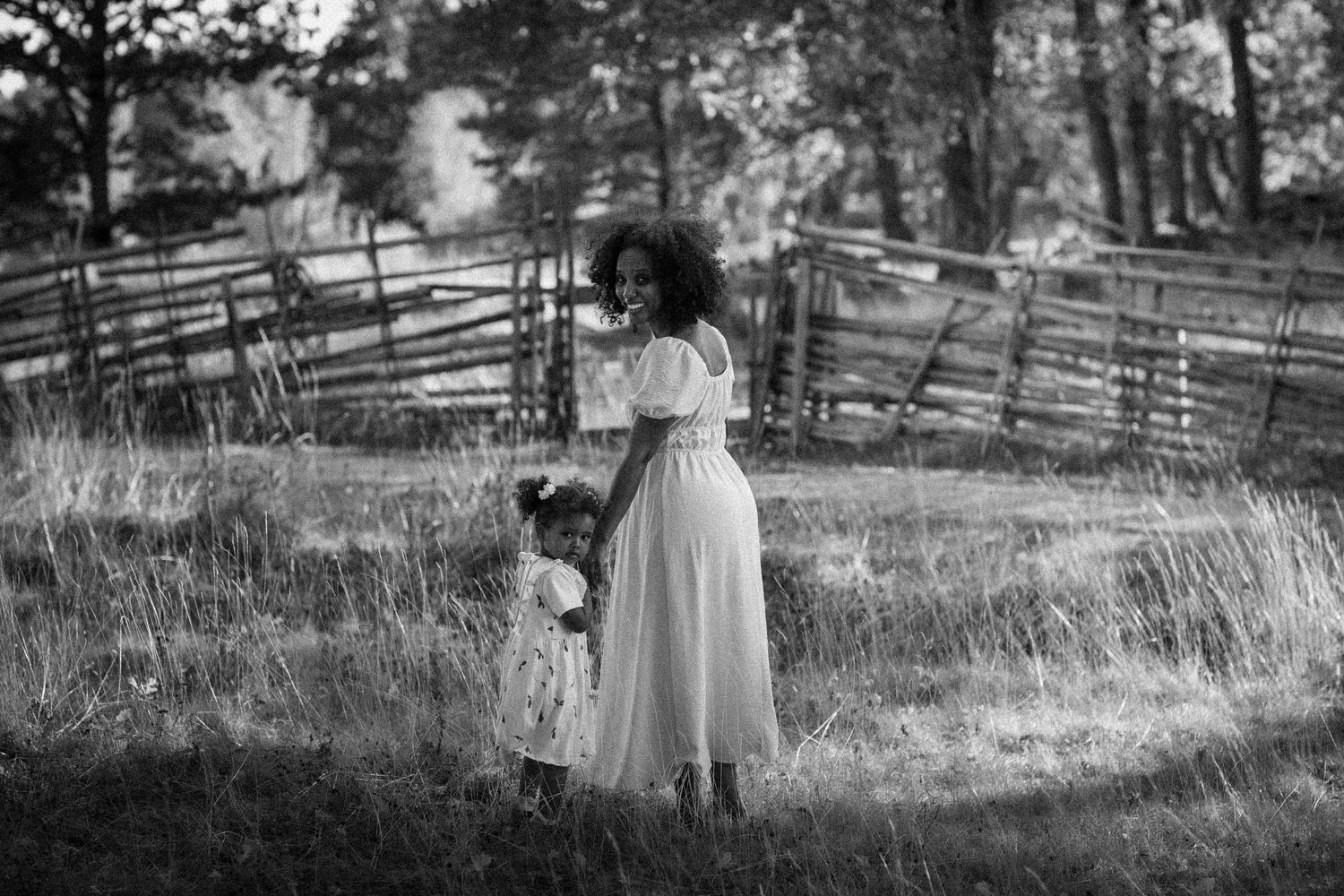 Black and white photo of a mother and child standing in a field near a wooden fence.