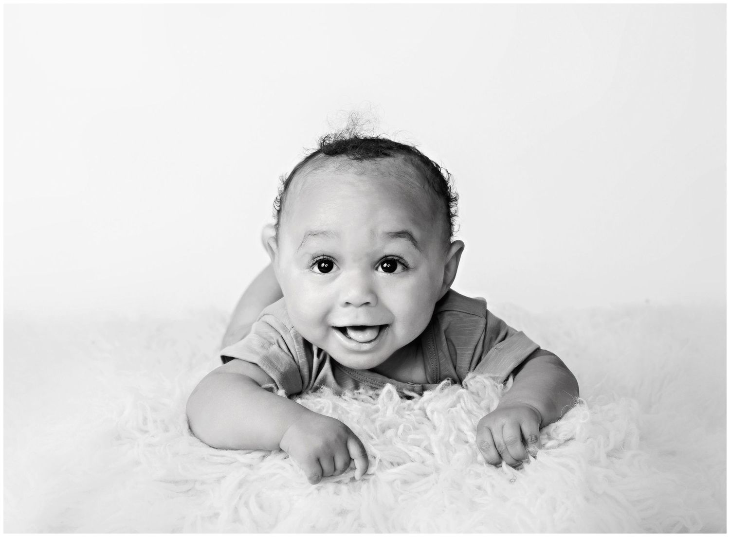 Tummy time portrait in black and white of smiling 3 month old baby boy posed on fluffy rug.