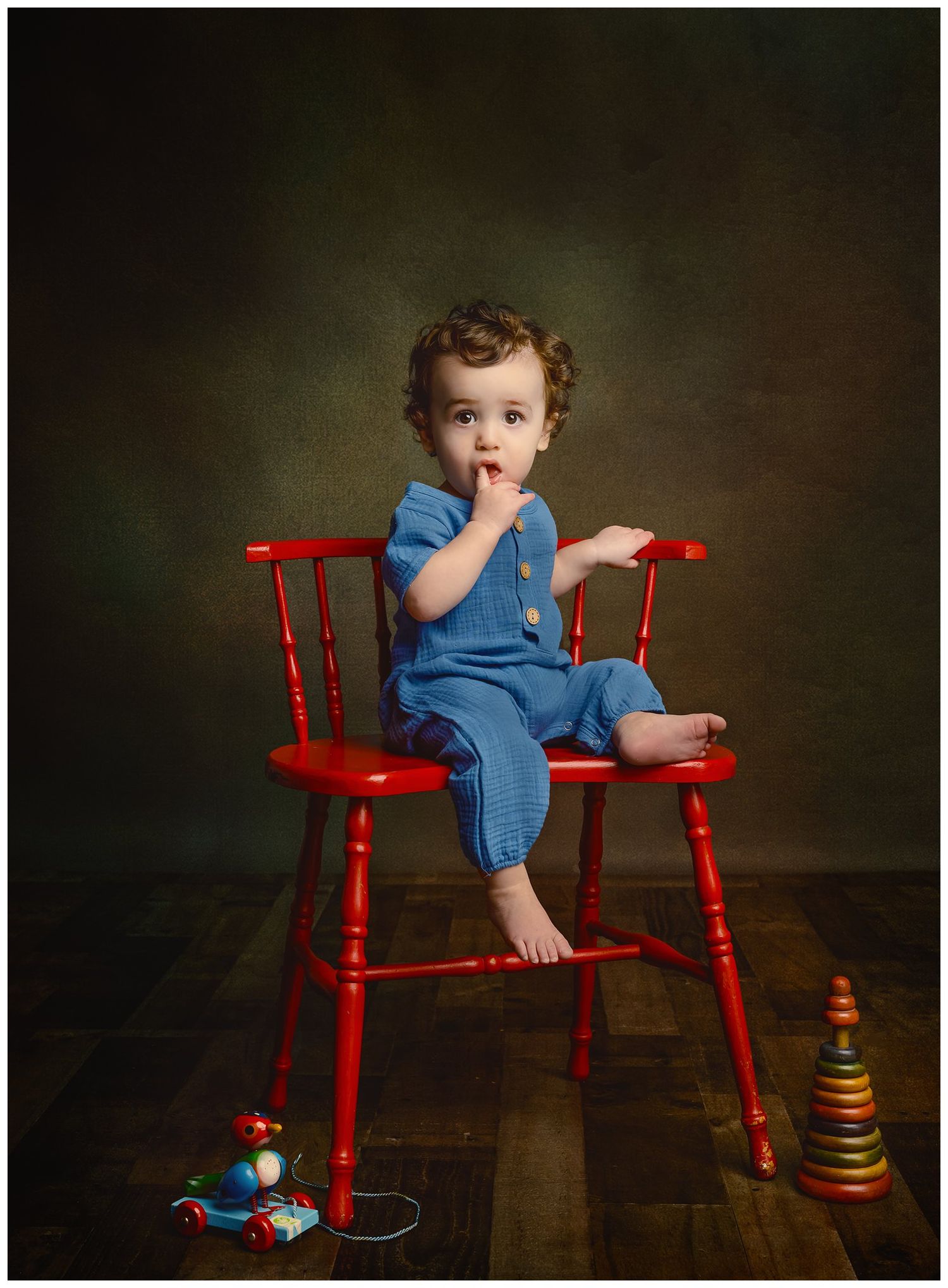 Portrait of 9 month old boy posed on a red spindle chair with vintage toys.  He is wearing a blue jumpsuit and has curly hair.  Austin Texas