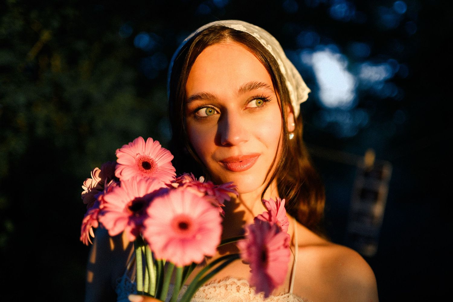 Young woman holding pink gerbera daisies in warm golden light, wearing a white headscarf outdoors at dusk.