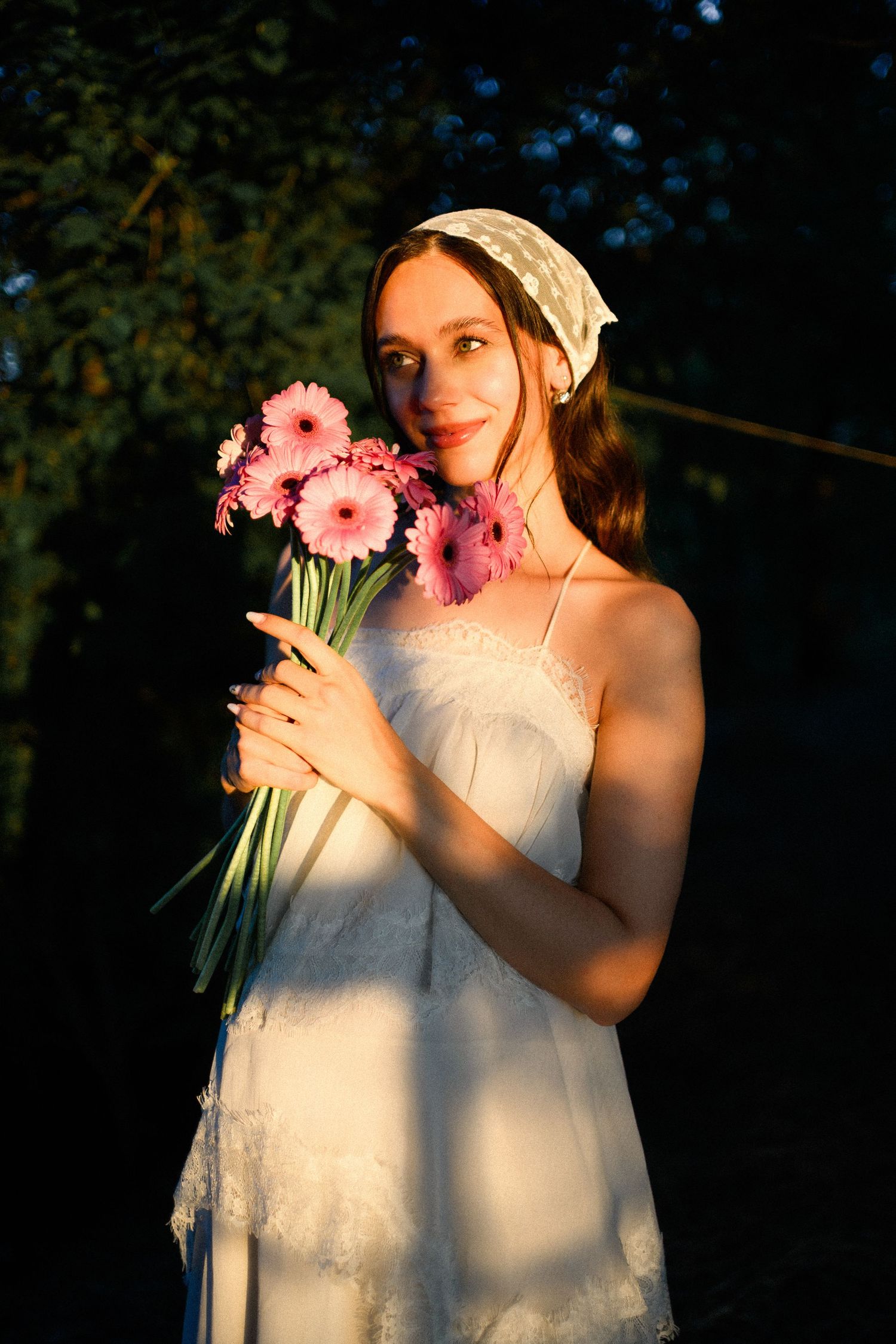 Young woman in white dress and headscarf holds pink gerbera daisies in golden sunset light outdoors.
