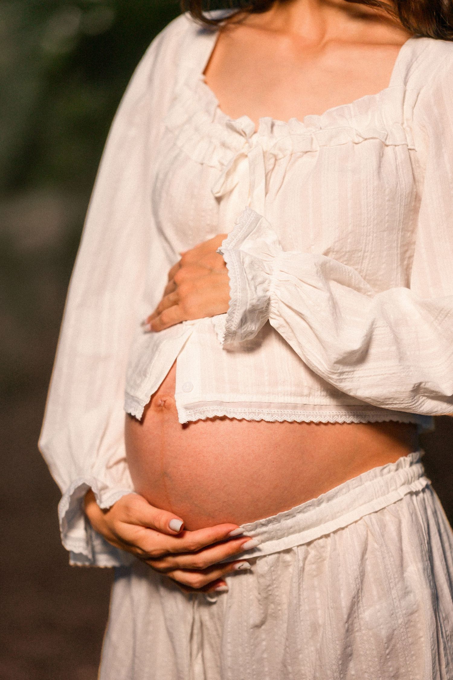 Pregnant woman in white boho dress gently cradling her baby bump outdoors in soft natural light.