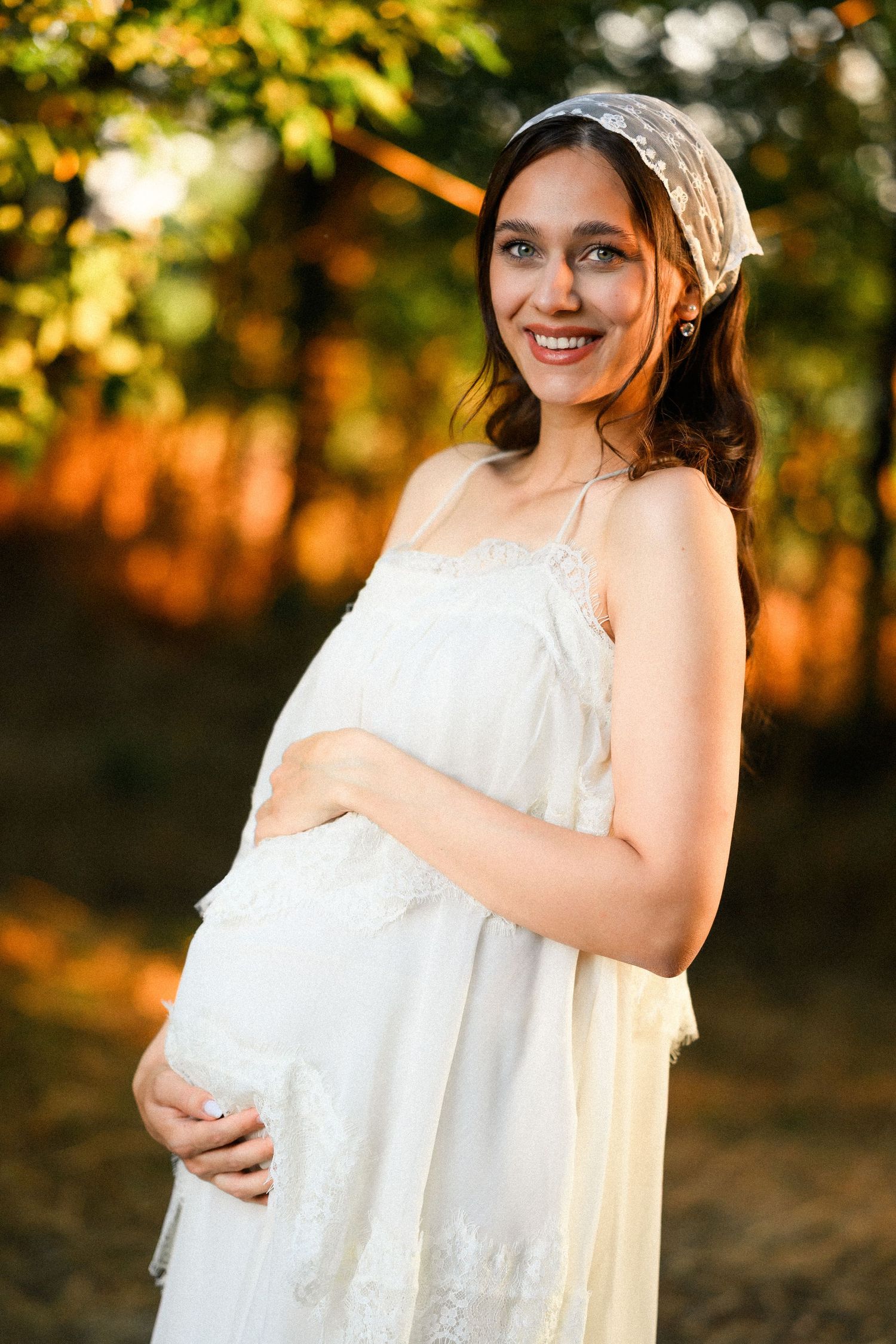 Smiling pregnant woman in white lace dress holding baby bump outdoors in golden sunlight.