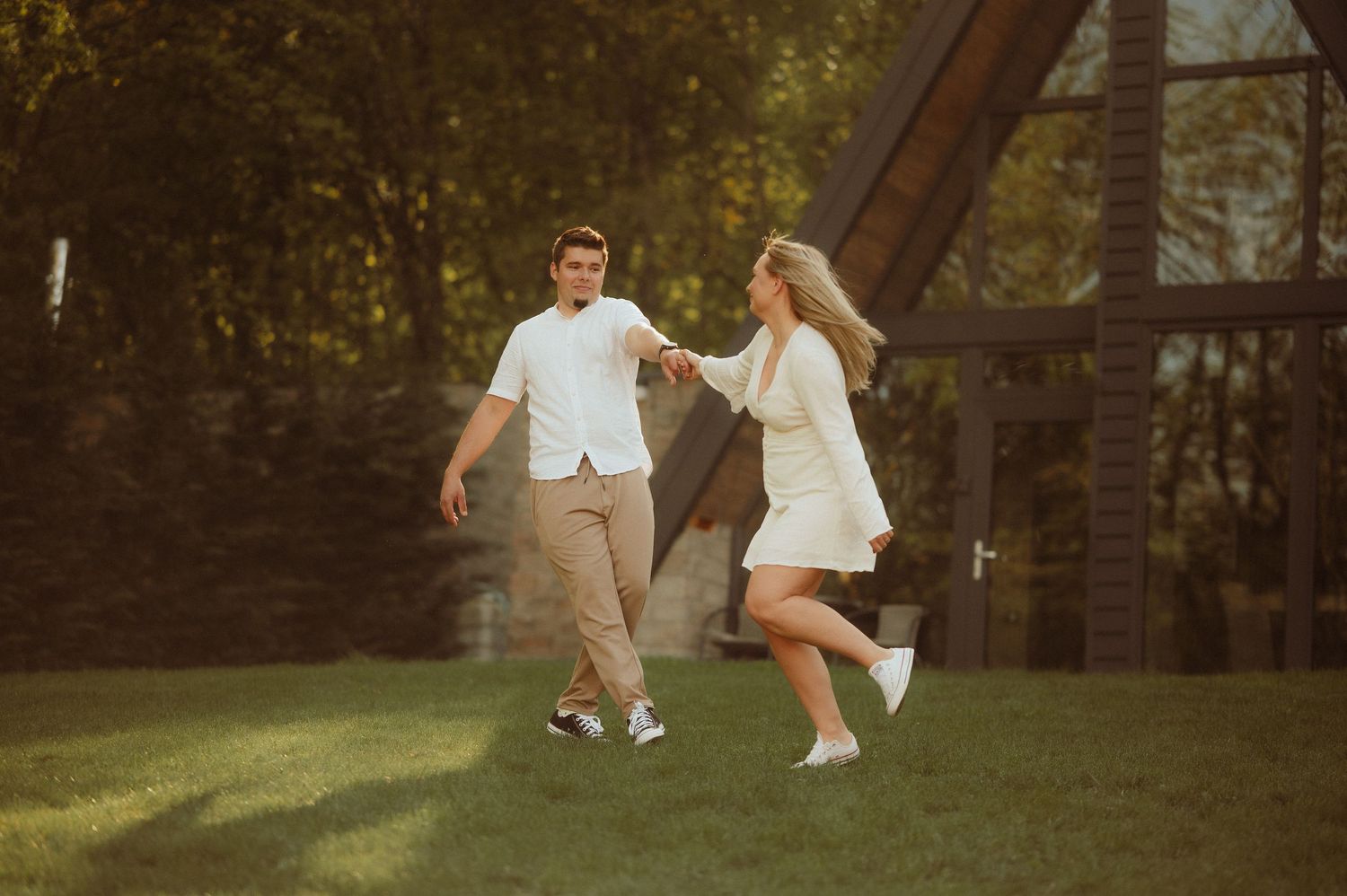 Couple playfully dancing together on green lawn near a wooden A-frame cabin in warm sunlight.