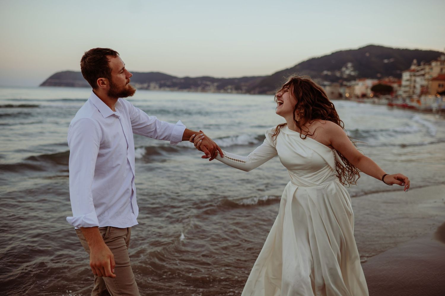 Happy couple holding hands and laughing on a beach at dusk by the sea.