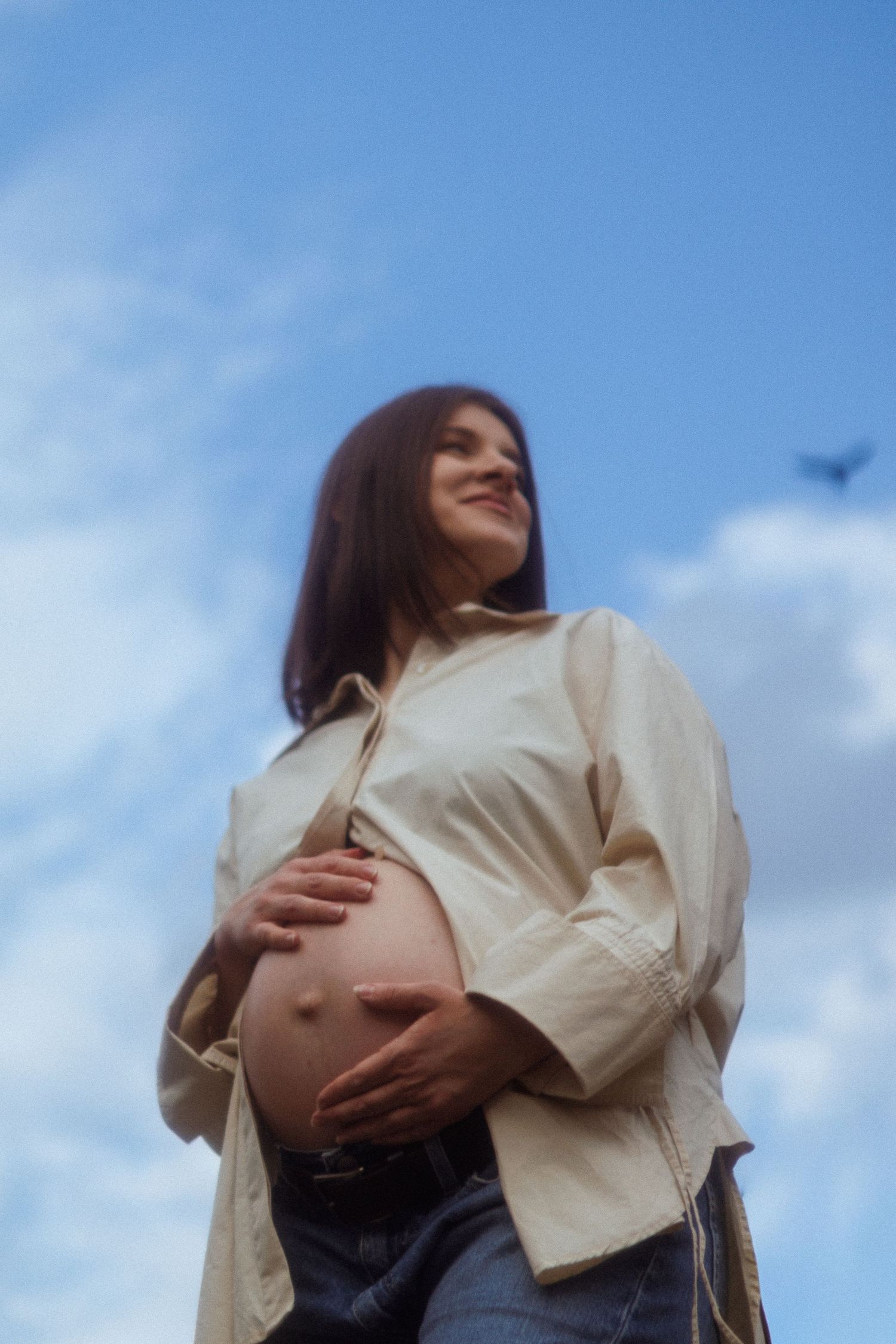 Pregnant woman in beige shirt holding baby bump against blue sky with clouds.