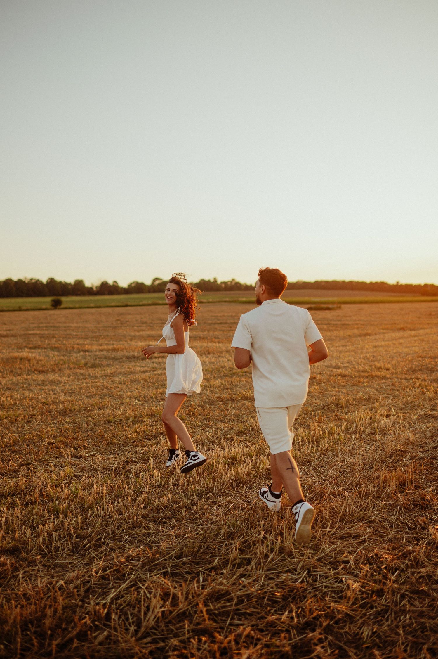 Couple in white outfits running through golden wheat field at sunset.