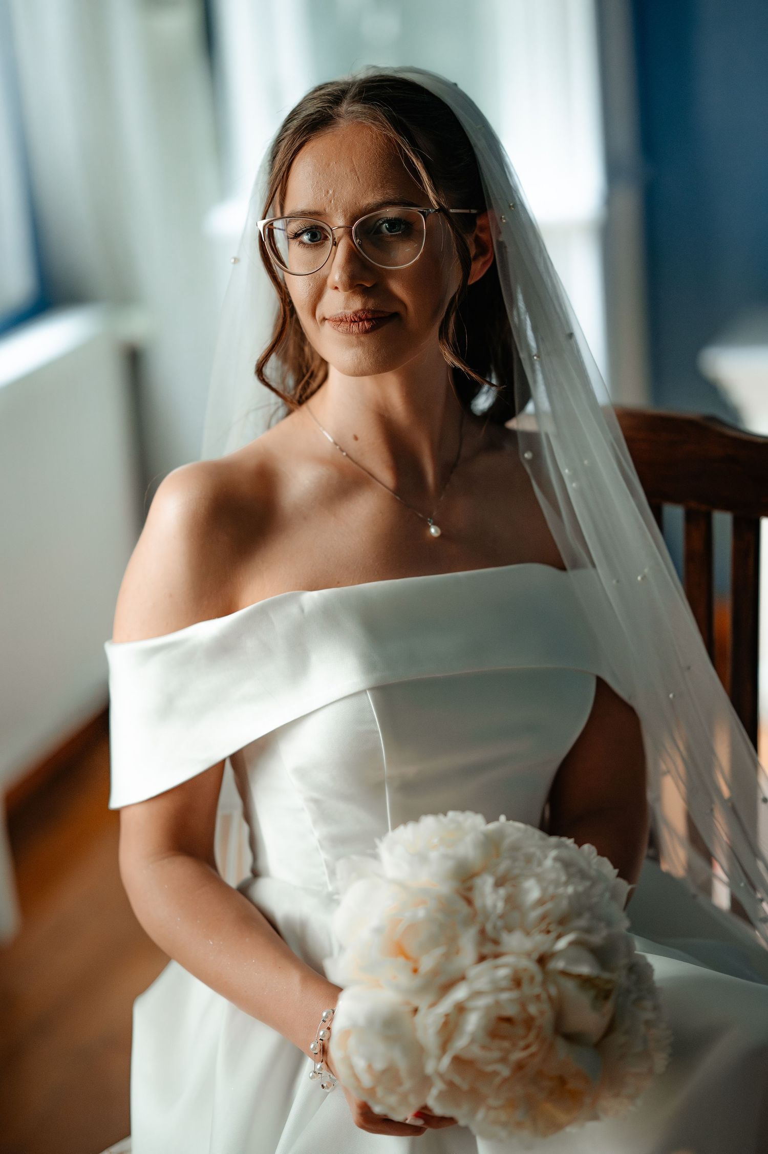 Bride in off-shoulder white gown, veil, and glasses holds white peony bouquet indoors.