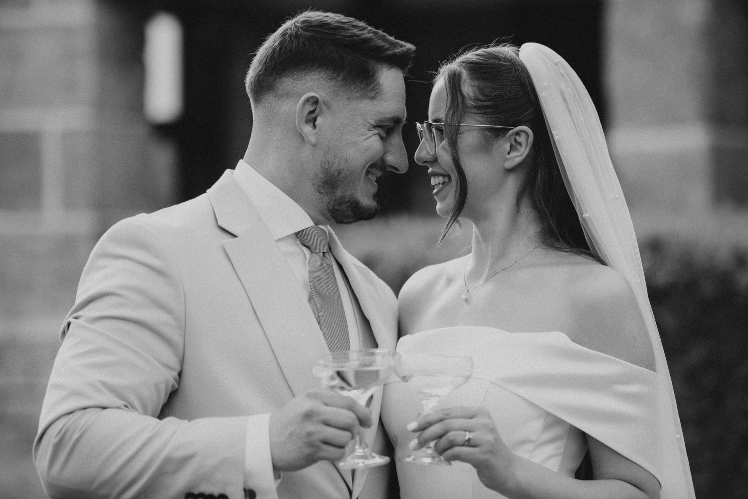Black and white portrait of smiling bride and groom touching foreheads while holding champagne glasses.