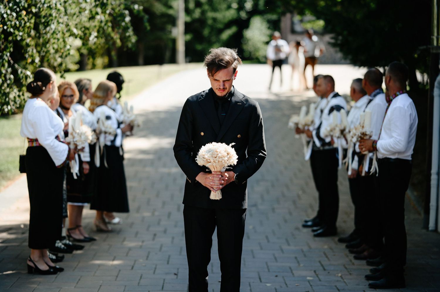 Groom in black military suit holds white bouquet while wedding party lines path.