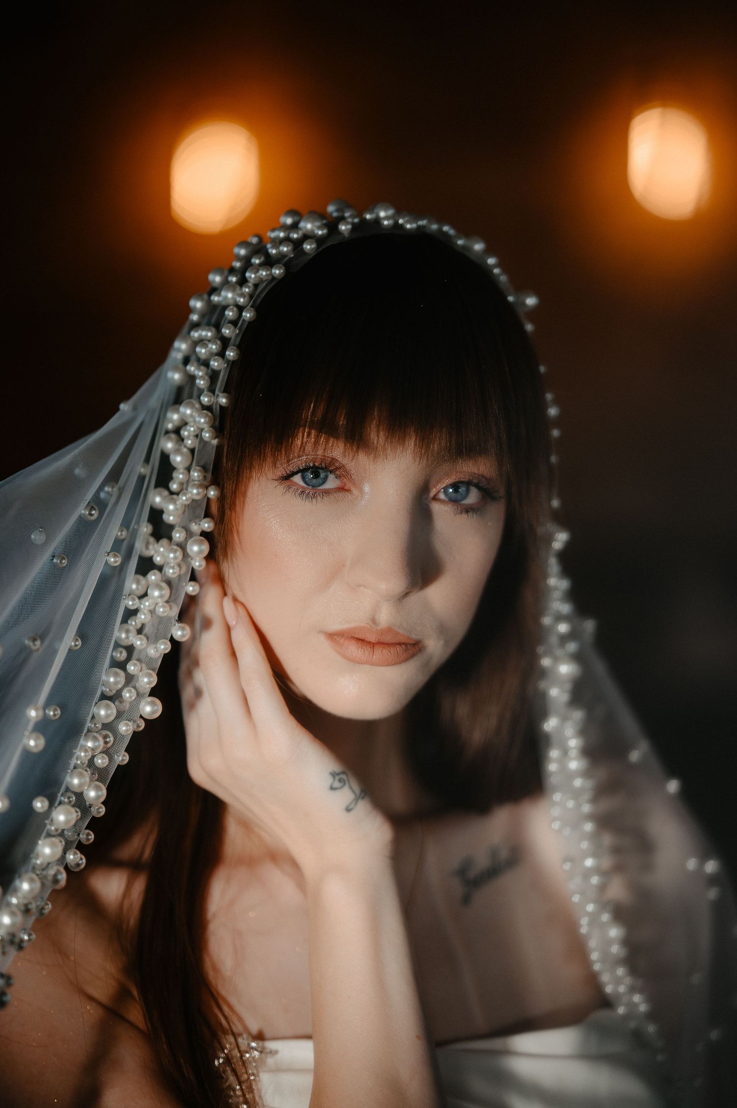 Beautiful bride with blue eyes wearing a pearl-trimmed veil gently touches her face in warm light.