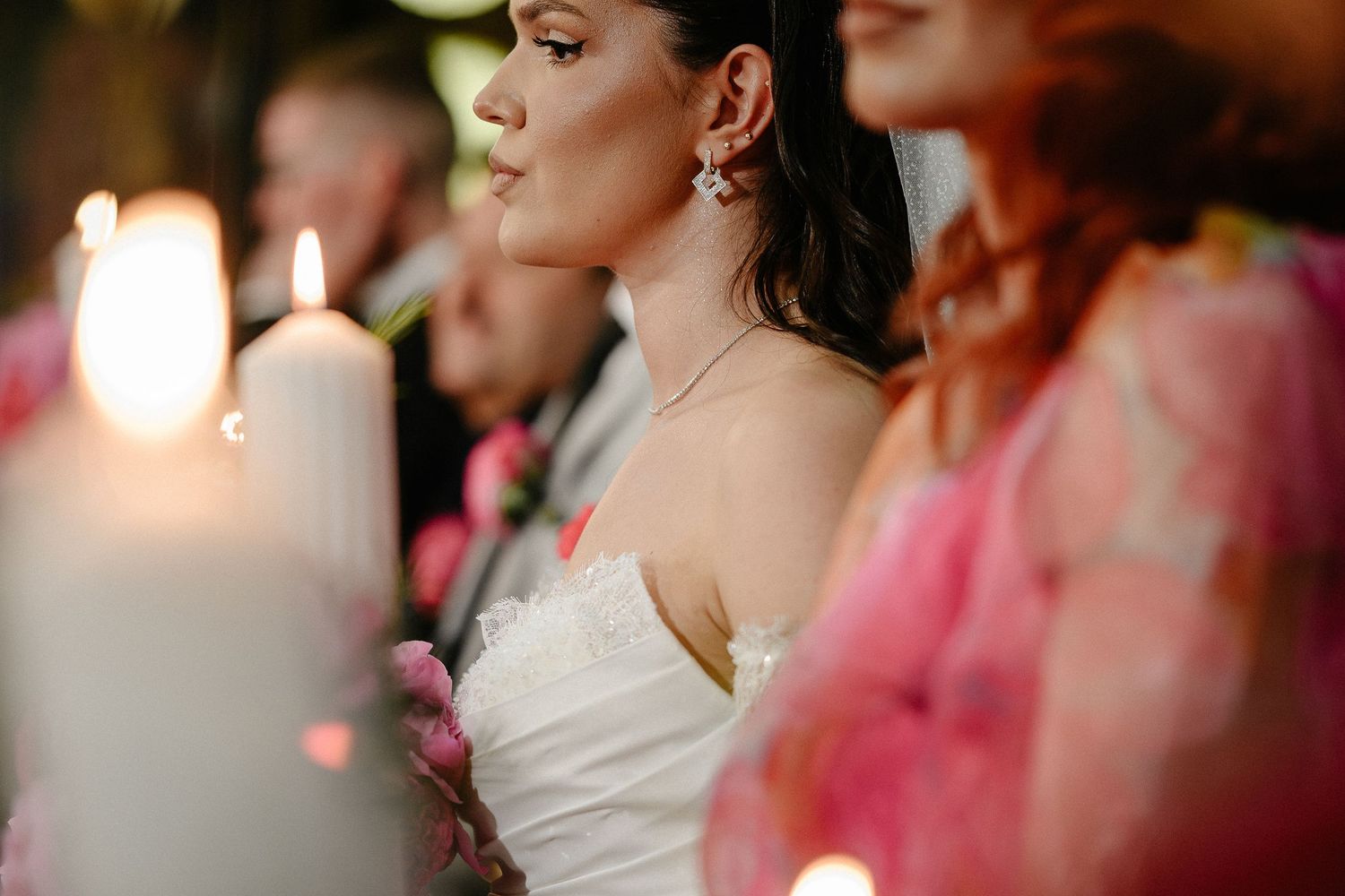 Bride in white strapless gown gazing peacefully beside glowing candles and pink flowers at her wedding.