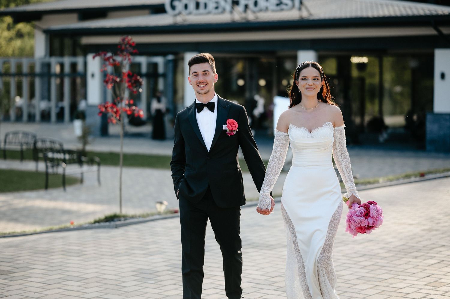 Wedding couple holding hands and walking toward camera outside Golden Forest, groom in black tuxedo with pink boutonniere.