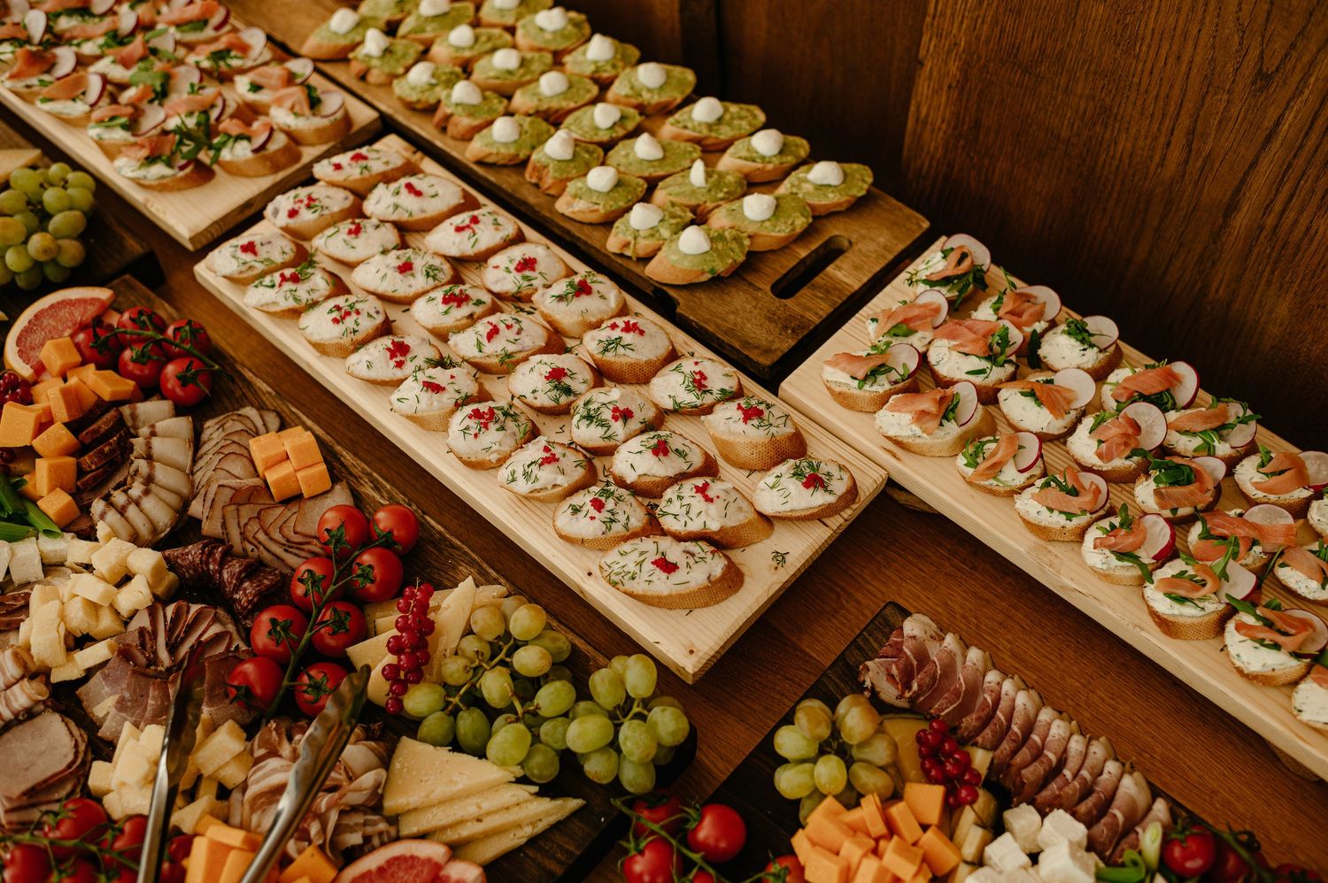 Elegant catering spread with appetizers on wooden boards surrounded by fresh fruits, cheeses, and charcuterie.
