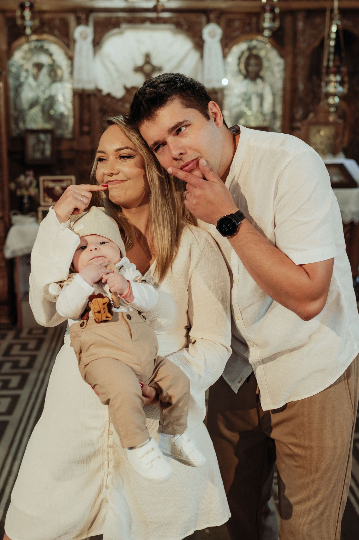 Young parents making funny faces while holding their baby during an Orthodox church baptism.