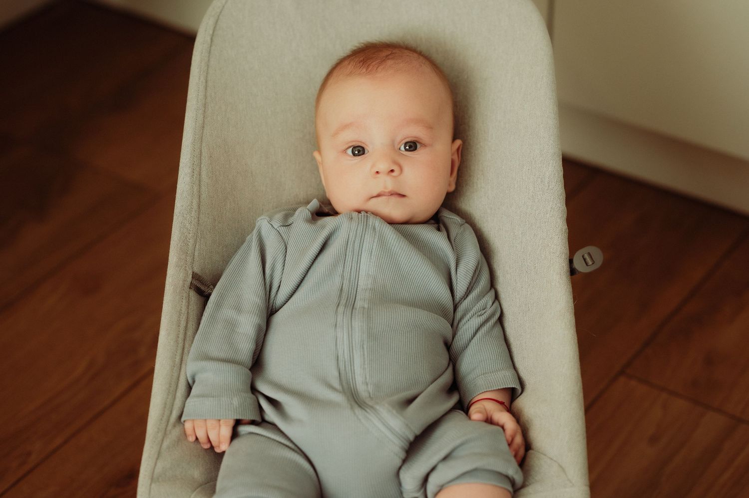 Adorable baby in sage green zip-up onesie lying in a soft infant bouncer chair.