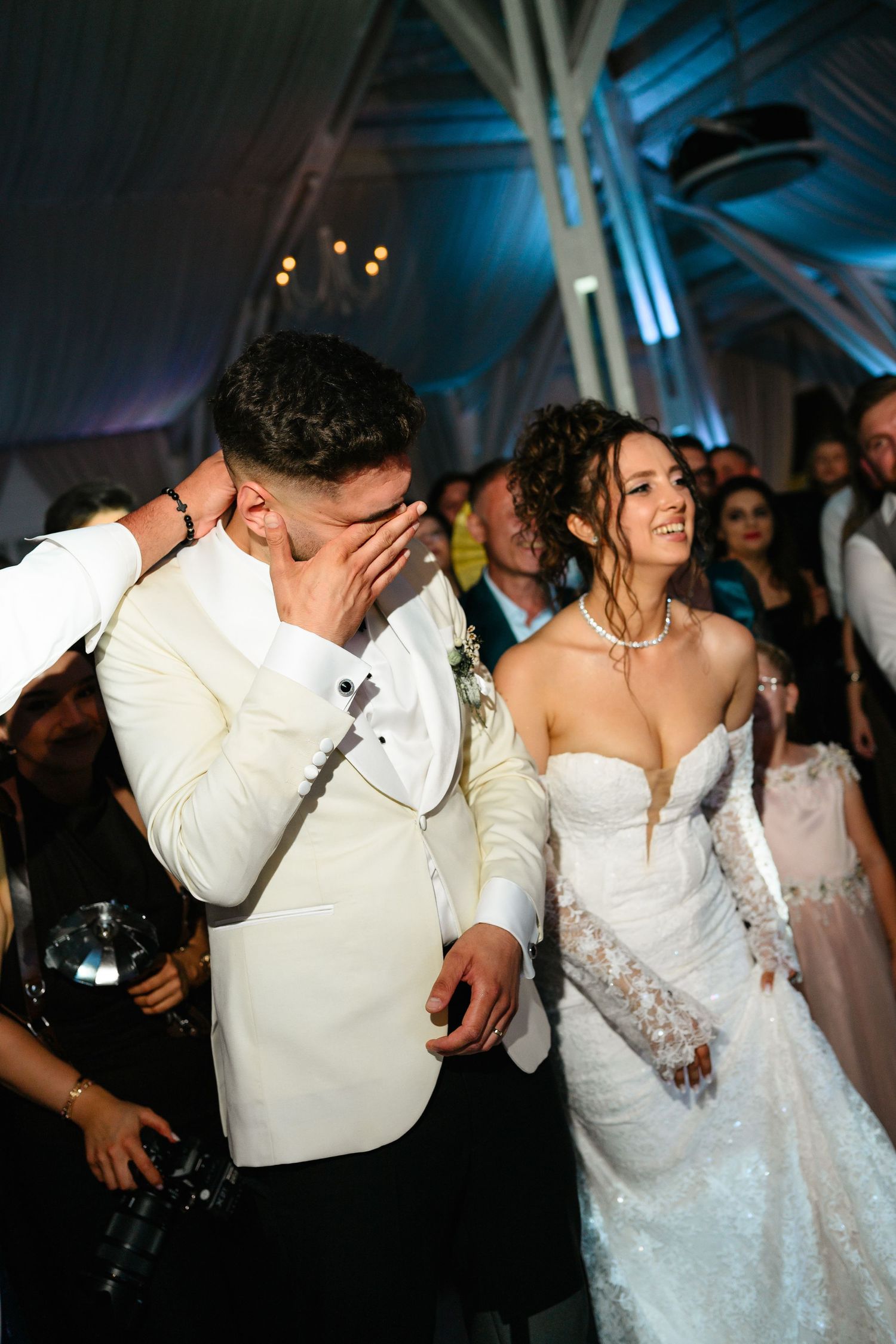 Emotional groom covering his face while laughing beside smiling bride in white lace gown during wedding reception.